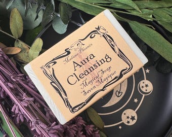 May include: A rectangular bar of Aura Cleansing Magical Soap with a tan label featuring black decorative text and a floral border. The soap is on a black plate with moon and star designs, surrounded by dried lavender and greenery.