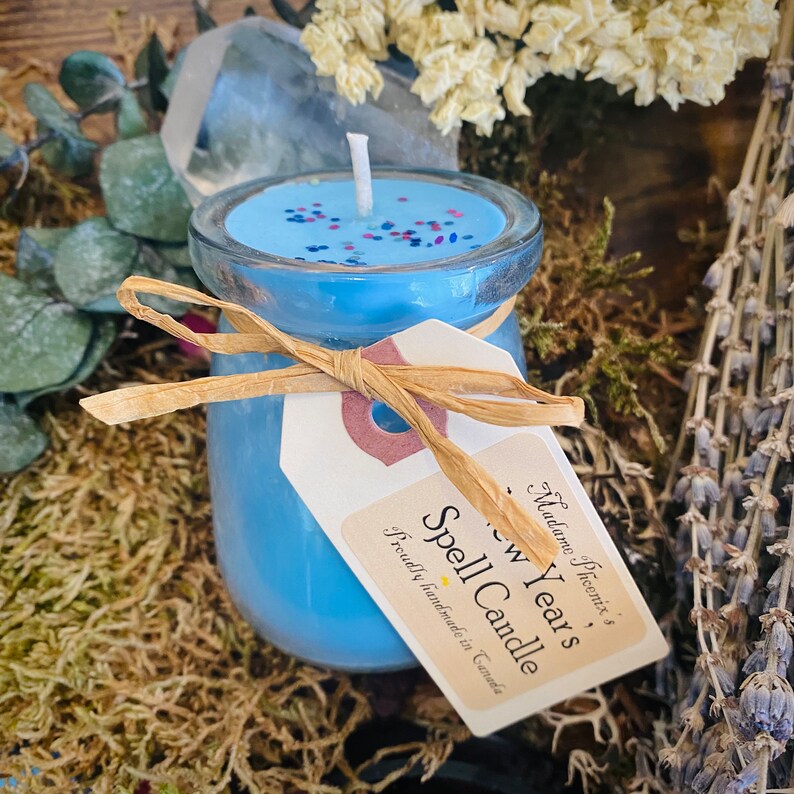 May include: A blue glass jar candle with a white wick and blue, red, and silver glitter. The candle is labelled "New Year's Spell Candle" and "Proudly handmade in Canada".
