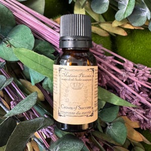 May include: A small amber glass bottle with a black screw-top cap and a beige rectangular label. The label features the text Madame Phoenix, magical oil, and Crown of Success, along with a crown illustration and decorative border elements.