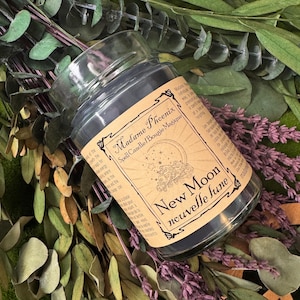 Könnte beinhalten: Eine Glas-Kerze mit dunkelgrauem Wachs, beschriftet mit "Madame Phoenix Spell Candle". Das Etikett zeigt den Text "New Moon nouvelle lune" und ein Mondsichel-Design. Die Kerze ist von getrocknetem Grün und Lavendel umgeben.