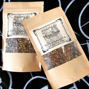 May include: Two brown paper bags with clear plastic windows showing loose leaf tea blends. The bags are labeled "Madame Pheoms's Moon Time" and "Madame Pheoms's Moon" with the text "regles de lune" and "Tea Blend/melange du the" in black and white.