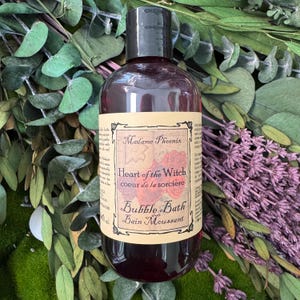 Peut inclure: Une bouteille de bain moussant rouge foncé avec un bouchon noir, étiquetée "Heart of the Witch" et "Bubble Bath". L'étiquette présente un motif floral et la marque "Madame Phoenix". La bouteille est entourée de feuillage vert et de fleurs violettes.