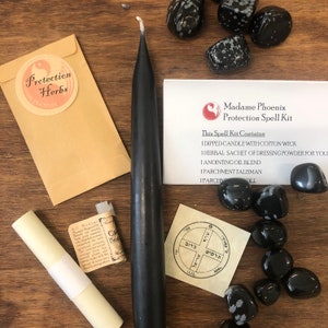 May include: A black candle, a small brown envelope with the words "Protection Herbs" and a symbol, a small white roll of parchment, a small white card with Hebrew text and a symbol, and a collection of black and white speckled stones on a wooden surface. The card also has the text "Madame Phoenix Protection Spell Kit" and "This Spell Kit Contains 1 Dipped Candle with Cotton Wick 1 Herbal Sachet of Dressing Powder for You 1 Anointing Oil Blend 1 Parchment Talisman 1 Parchment Roll"