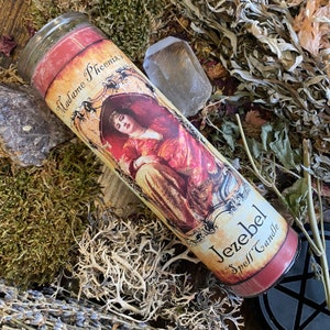Può includere: Una candela cilindrica in vetro con un'etichetta recante il testo Madame Phoenix e Jezebel Spell Candle. L'etichetta mostra l'illustrazione di una persona con abiti rossi e dorati. La candela è riempita di cera rossa e ha un coperchio in vetro.