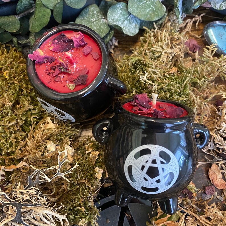 May include: Two black ceramic cauldron candles with red wax and dried rose petals. The candles feature a white pentagram design.