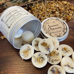 May include: A silver tin with a white label that reads "Sweet Dreams Wax Drops" and a floral design. The tin is open and wax melts are spilling out. The wax melts are white and have dried lavender and chamomile flowers on top.