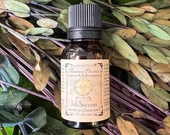 May include: A small amber glass bottle with a black screw-top cap, featuring a beige rectangular label with the text Madame Phoenix magical oil, Job Success, and a circular emblem. The bottle is surrounded by dried green and brown eucalyptus leaves.