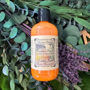 May include: An orange bubble bath bottle with a black cap, labeled "Pumpkin Patch" and "Bubble Bath." The bottle is surrounded by green eucalyptus leaves and purple lavender sprigs, creating a natural, spa-like aesthetic.
