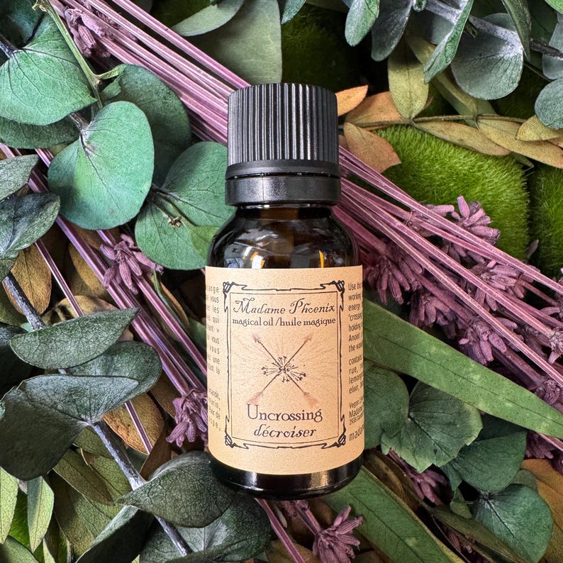 Pu&ograve; includere: Una piccola bottiglia in vetro ambrato con tappo nero a vite e un'etichetta di carta color cuoio. L'etichetta riporta il testo Madame Phoenix magical oil, Uncrossing e un disegno di due frecce incrociate. La bottiglia &egrave; circondata da foglie di eucalipto.