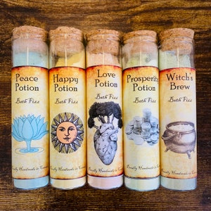 Peut inclure: Cinq bouteilles en verre avec des bouchons en liège et des étiquettes. Les étiquettes sont décorées d'illustrations et de texte. Le texte sur les étiquettes indique "Potion de paix", "Potion de bonheur", "Potion d'amour", "Potion de prospérité" et "Brassée de sorcière". Toutes les bouteilles sont étiquetées "Bain moussant".