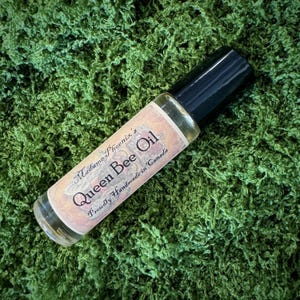 Può includere: Un flacone roll-on di vetro trasparente di Queen Bee Oil con tappo nero, appoggiato su un letto di muschio verde. L'etichetta presenta il testo "Queen Bee Oil" e "Proudly Handmade in Canada".