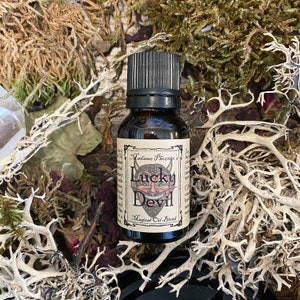 May include: A small brown glass bottle with a black cap. The label reads "Madame Phoenix's Lucky Devil Magical Oil Blend". The label features a cartoon image of a red devil with horns and a mischievous grin.