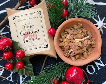 May include: A brown paper pouch with the text "Madame Phoenix's Yule Garland Incense" and the words "Proudly Hand-Made in Canada". The pouch is sitting on a black and white surface with red berries and green pine branches. A small terracotta bowl filled with brown incense is also visible.