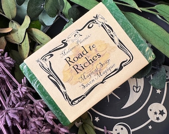 Può includere: Una saponetta verde con un'etichetta che recita "Madame Phoenix Road to Riches Magical Soap Savon Magique". Il sapone è circondato da lavanda secca e verde, con un piatto decorativo nero sullo sfondo.