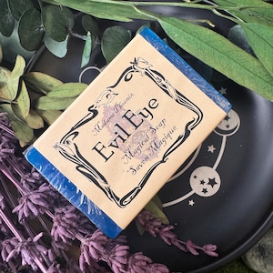 May include: A rectangular bar of soap with the words "Evil Eye Magical Soap Savon Magique" printed on the label. The label is tan with a black decorative border. The soap is on a black plate with a celestial design, surrounded by lavender and greenery.