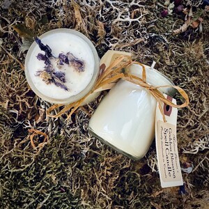 May include: Two white soy candles in glass jars. One jar contains dried purple flowers. The other jar has a label that reads "Goddess Blessing Spell Candle" and "Handcrafted in Canada".