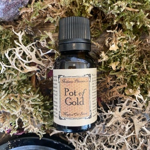 May include: A small brown glass bottle with a black cap, labelled "Madame Phoenix's Pot of Gold Magical Oil Blend". The bottle is sitting on a bed of green moss.