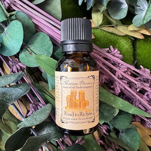 May include: A small amber glass bottle with a black cap and a tan label. The label features an illustration of stacked gold coins and the text Madame Phoenix magical oil, Road to Riches. The bottle is surrounded by green leaves and purple dried flowers.