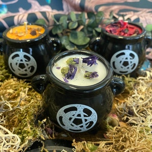 May include: Three black cauldron-shaped candle holders with white pentagram designs. The candles are topped with dried flowers and herbs.