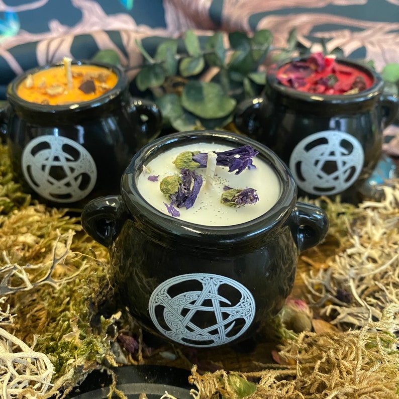 May include: Three black cauldron-shaped candle holders with white pentagram designs. The candles are topped with dried flowers and herbs.