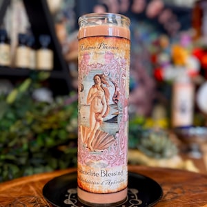 May include: A tall cylindrical glass candle filled with peach-coloured wax. The label features the text Madame Phoenix and Aphrodite Blessing, with an illustration of a figure standing in a seashell on a beach, framed by ornate pink floral borders.