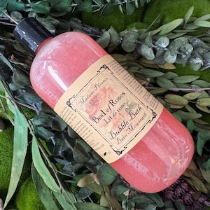 May include: A clear plastic bottle filled with pink bubble bath liquid, topped with a black cap. The label features the text Madame Phoenix, Bed of Roses, Lit de rose, and Bubble Bath, Bain Moussant, with a rose illustration and descriptive text.