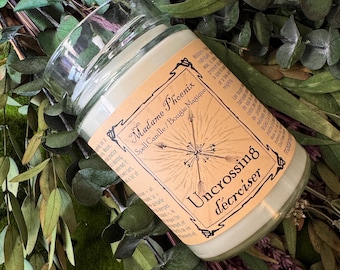Könnte beinhalten: Eine Kerze im klaren Glasgefäß mit hellfarbigem Wachs. Das beigefarbene Etikett trägt den schwarzen Text Madame Phoenix Spell Candle / Bougie Magique und Uncrossing / décroiser sowie eine zentrale Grafik mit nach außen gerichteten Pfeilen.