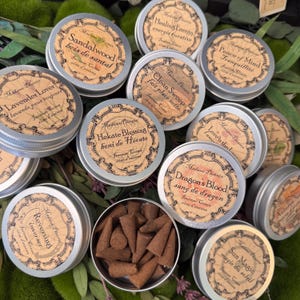 May include: A collection of round silver metal tins filled with incense cones, each featuring a beige label with black text. Labels include names like Sandalwood, Healing Energy, Peace of Mind, Clean Sweep, Hekate Blessing, and Dragon's Blood.