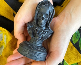 Può includere: Portacandele nero con una figura incappucciata con un teschio. La base del portacandele è rotonda e ha la scritta "Santa Muerte".
