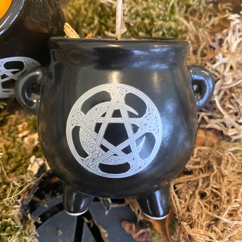 May include: Black cauldron candle holder with a white pentagram and moon design. The cauldron has three legs and is sitting on a bed of moss.