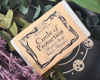 Könnte beinhalten: Ein Seifenstück in einer beigefarbenen Verpackung mit schwarzem Text, der "Circle of Protection Magical Soap Savon Magique" lautet. Die Seife liegt neben getrocknetem Lavendel und Grünzeug auf einer dunklen Oberfläche.