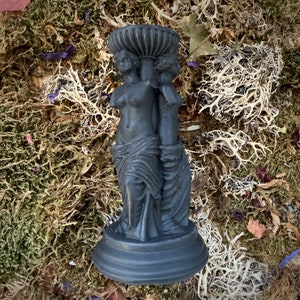 May include: Black candle holder featuring two female figures sculpted in a classical style. The holder has a round base and a decorative top.