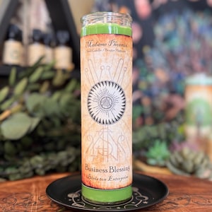 May include: A tall green pillar candle in a clear glass jar with a tan label. The label features the text Madame Phoenix, Spell Candle, and Business Blessing, along with a central circular graphic containing a triangle and radiating lines.