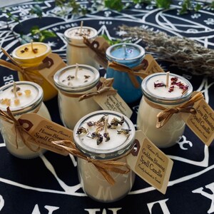 May include: An assortment of small glass jar candles with various scents and dried flowers on top. The candles are labelled with the text "Goddess Blessing Spell Candle", "House Blessing Spell Candle", "Love Blessing Spell Candle" and "Romance Blessing Spell Candle".