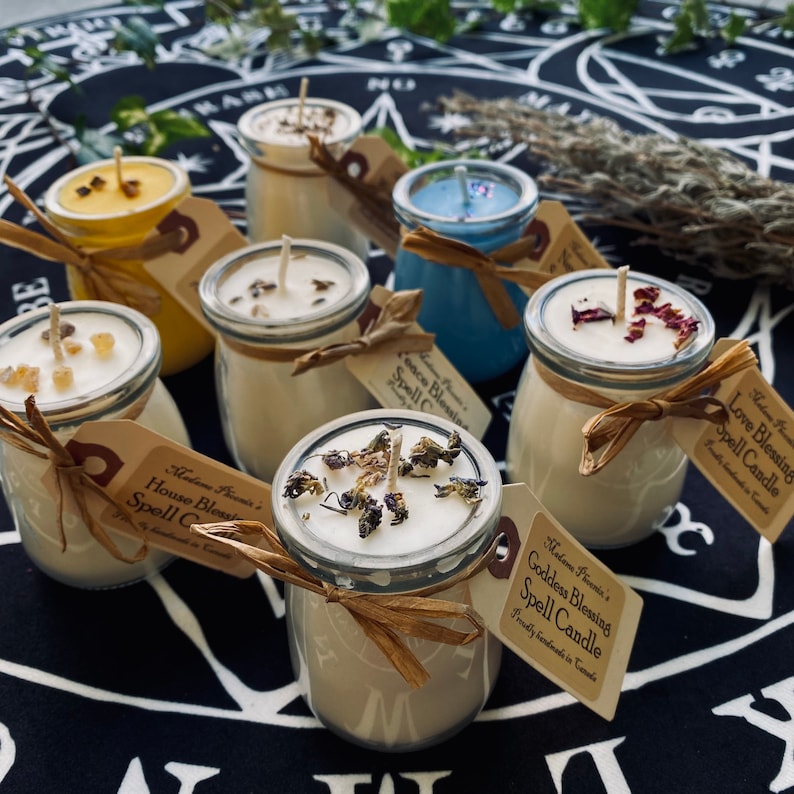 May include: An assortment of small glass jar candles with various scents and dried flowers on top. The candles are labelled with the text "Goddess Blessing Spell Candle", "House Blessing Spell Candle", "Love Blessing Spell Candle" and "Romance Blessing Spell Candle".