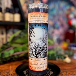May include: A tall cylindrical glass candle filled with dark wax. The label features the text Madame Phoenix Spell Candle, an illustration of a full moon behind bare tree branches, and the text Shadows & Moonlight at the bottom of the label.