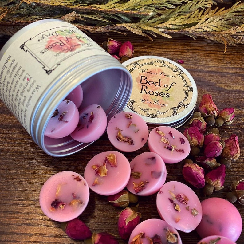 May include: A silver tin with a label that reads "Madame Phoenix Bed of Roses Wax Drops" is open and spilling out pink wax melts with dried rose petals. The wax melts are scattered on a wooden surface with dried rose buds.