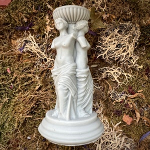 May include: A white ceramic candle holder featuring two sculpted figures of women standing back to back. The figures are draped in flowing robes and have a classic, elegant style.