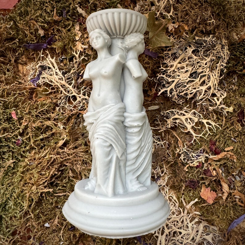 May include: A white ceramic candle holder featuring two sculpted figures of women standing back to back. The figures are draped in flowing robes and have a classic, elegant style.