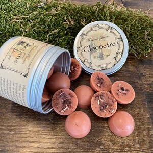 May include: A silver tin with a label that reads "Madame Phoenix Cleopatra Wax Drops". The tin is open and wax melts are spilling out. The wax melts are a light brown colour and have a speckled appearance.