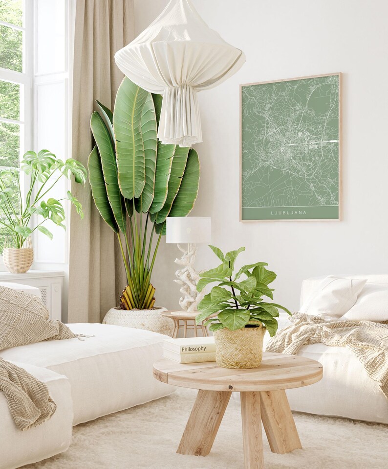 May include: A bright, airy living room with a large green plant, a framed map of Ljubljana, and a small wooden table. The room features white sofas, a woven basket, and a cream-colored rug. A white lamp and a decorative light fixture are also visible.