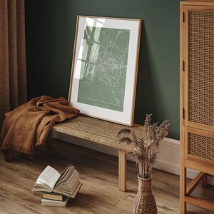 May include: A framed map print of Ljubljana in a light green hue, displayed on a woven bench. A brown throw blanket rests on the bench, and a stack of books sits on the wooden floor. A wicker vase holds dried plants.