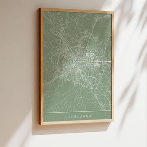 May include: A framed map print of Ljubljana, Slovenia, in a minimalist style. The map features white lines on a muted green background, with the city name at the bottom. The frame is a light wood color.