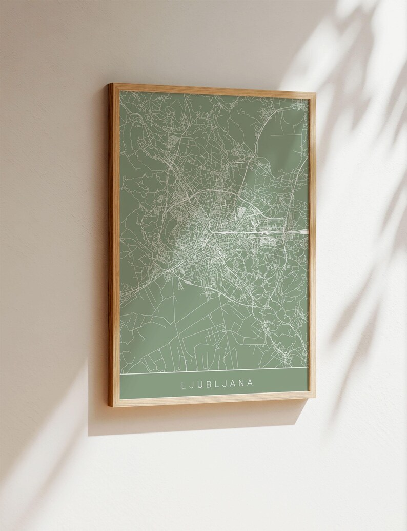 May include: A framed map print of Ljubljana, Slovenia, in a minimalist style. The map features white lines on a muted green background, with the city name at the bottom. The frame is a light wood color.