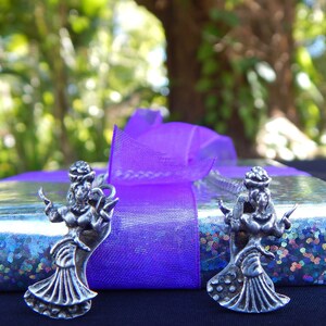 May include: Two silver charm pendants in the shape of a woman dancing, with a purple ribbon in the background.