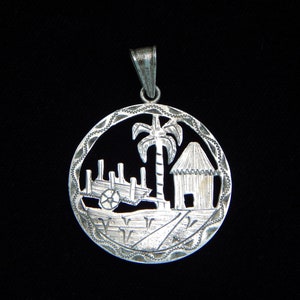 May include: Silver pendant featuring a cutout design of a palm tree, a small hut, and a cart with wheels. The pendant is circular and has a textured border.