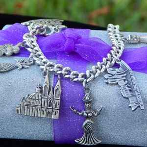 May include: A silver charm bracelet with multiple charms, including a cathedral, a mermaid, a hand, and a building. The bracelet is on a silver background with a purple ribbon.
