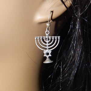 May include: Silver menorah earrings with a Star of David charm. The earrings are hanging from a person's ear.