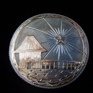 May include: A silver circular ornament with a detailed design of a star with rays, a small house, and a fence. The ornament has a textured border.
