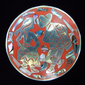 May include: A silver and abalone shell inlay dish with a red background. The dish features a stylized design of a man and woman dancing.